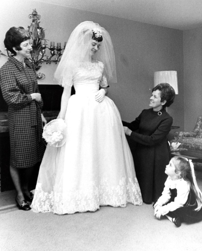 Black and white image of a standing bride looking down at a child seated on the floor. A crouched attendant adjusts her wedding gown, while another woman looks on.